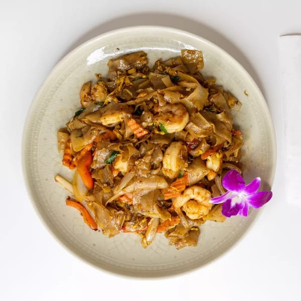 Pad See Ew with shrimp, carrots, and onions, garnished with a purple orchid and red chopsticks.