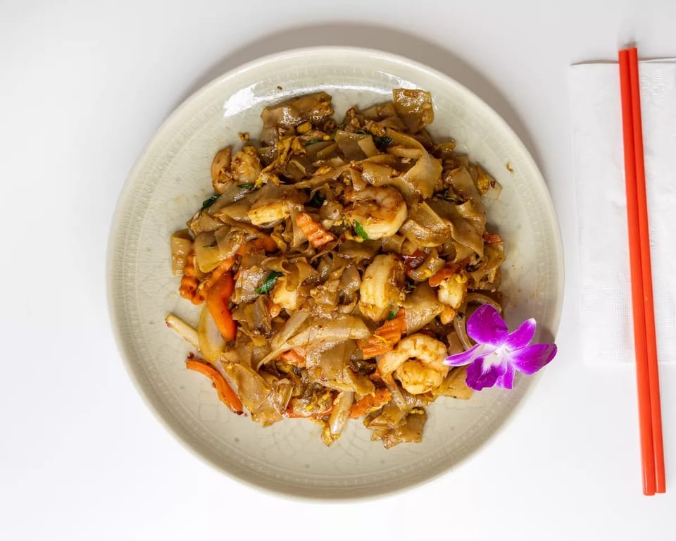 Pad See Ew with shrimp, carrots, and onions, garnished with a purple orchid and red chopsticks.