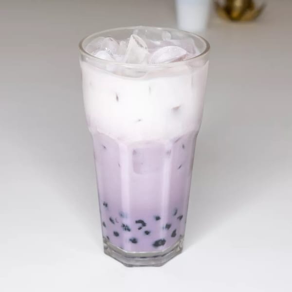 Tall glass of taro bubble tea with a creamy white top layer and ice on a white surface.