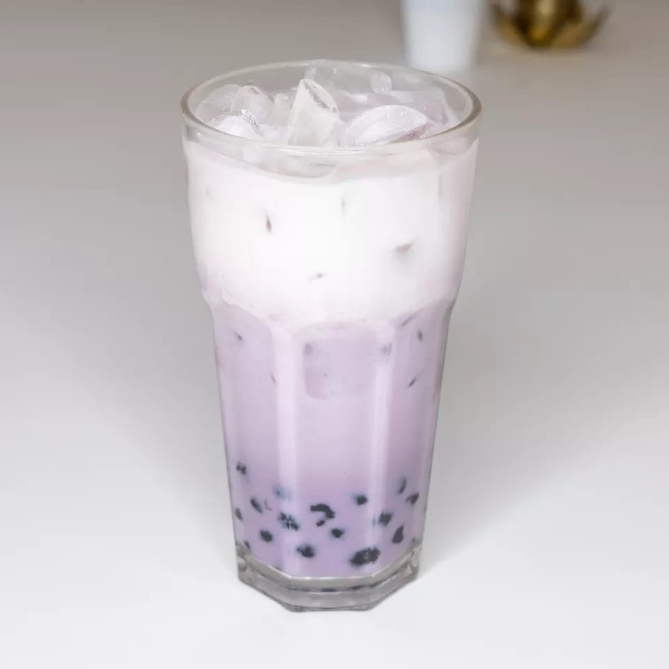Tall glass of taro bubble tea with a creamy white top layer and ice on a white surface.