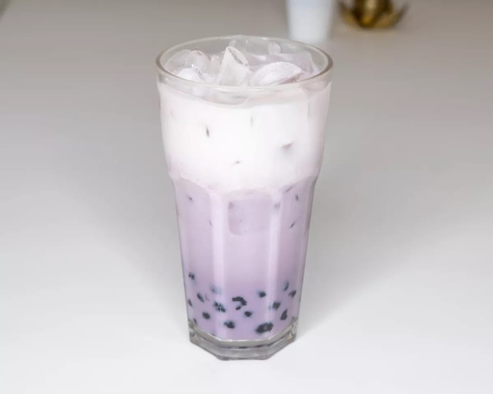 Tall glass of taro bubble tea with a creamy white top layer and ice on a white surface.