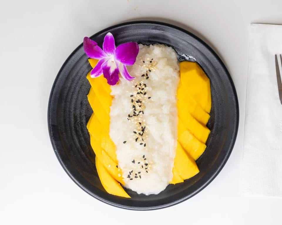 Mango sticky rice dessert with a purple orchid, black sesame, and white sesame seeds.