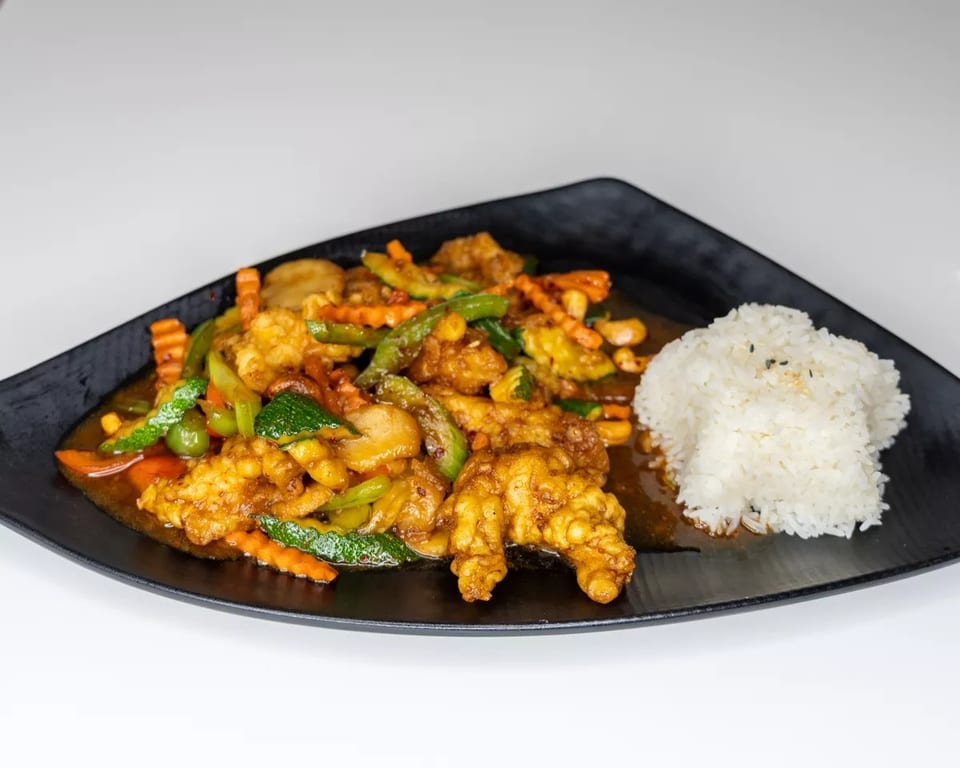 Crispy chicken stir-fry with colorful vegetables and white rice on a black plate.
