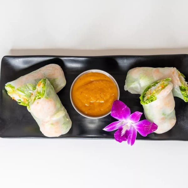 Four fresh spring rolls with shrimp and vegetables, a side of peanut sauce, and a purple orchid.