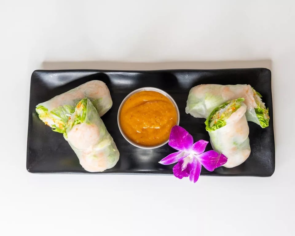 Four fresh spring rolls with shrimp and vegetables, a side of peanut sauce, and a purple orchid.